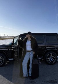 Femme portant un long manteau en fourrure noire, un haut blanc et un jean, debout à côté d'une porte ouverte d'un SUV noir par une journée ensoleillée.