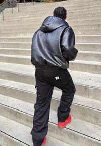 Czarna skórzana kurtka z kapturem, czarne spodnie typu cargo z logo Carhartt i czerwone obuwie; wchodzenie po betonowych schodach.