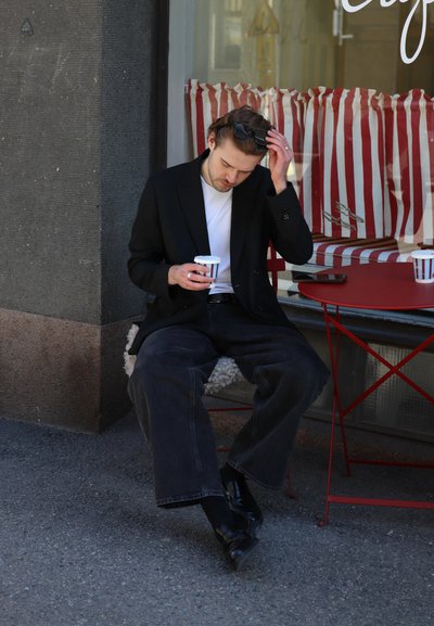 Blazer negro sobre una camisa blanca, pantalones oscuros de pierna ancha. Sosteniendo una taza a rayas, sentado cerca de una mesa de bistró roja. De fondo, cortinas a rayas.