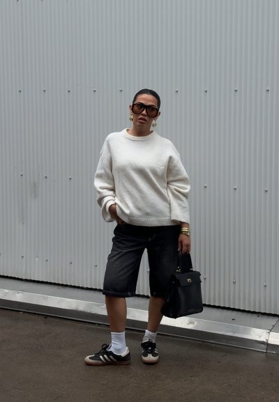 Suéter blanco texturizado, pantalones cortos oscuros tipo bermuda, bolso negro, zapatillas negras y blancas. Grandes gafas de sol y pendientes dorados completan el look.