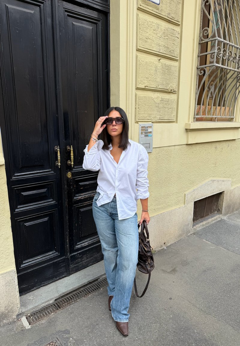 Mulher com camisa branca e jeans azuis a ajustar os óculos de sol, segurando uma bolsa castanha, a pé junto a uma porta preta e uma parede amarela na calçada.