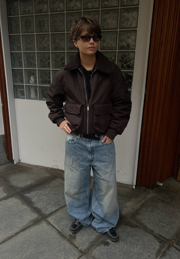 Jeune personne portant des lunettes de soleil foncées, une veste zippée noire, un jean large délavé et des chaussures noires, debout sur le trottoir près d'un mur en briques de verre.