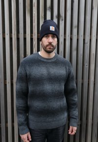 Pull gris avec un motif rayé texturé et des poignets côtelés. Porté avec un bonnet bleu marine comportant un petit patch logo. Fond composé de lattes en bois.