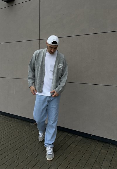 Cárdigan gris de punto con botones sobre una camiseta blanca, combinado con jeans azul claro y zapatillas grises, sobre un fondo de pared gris.