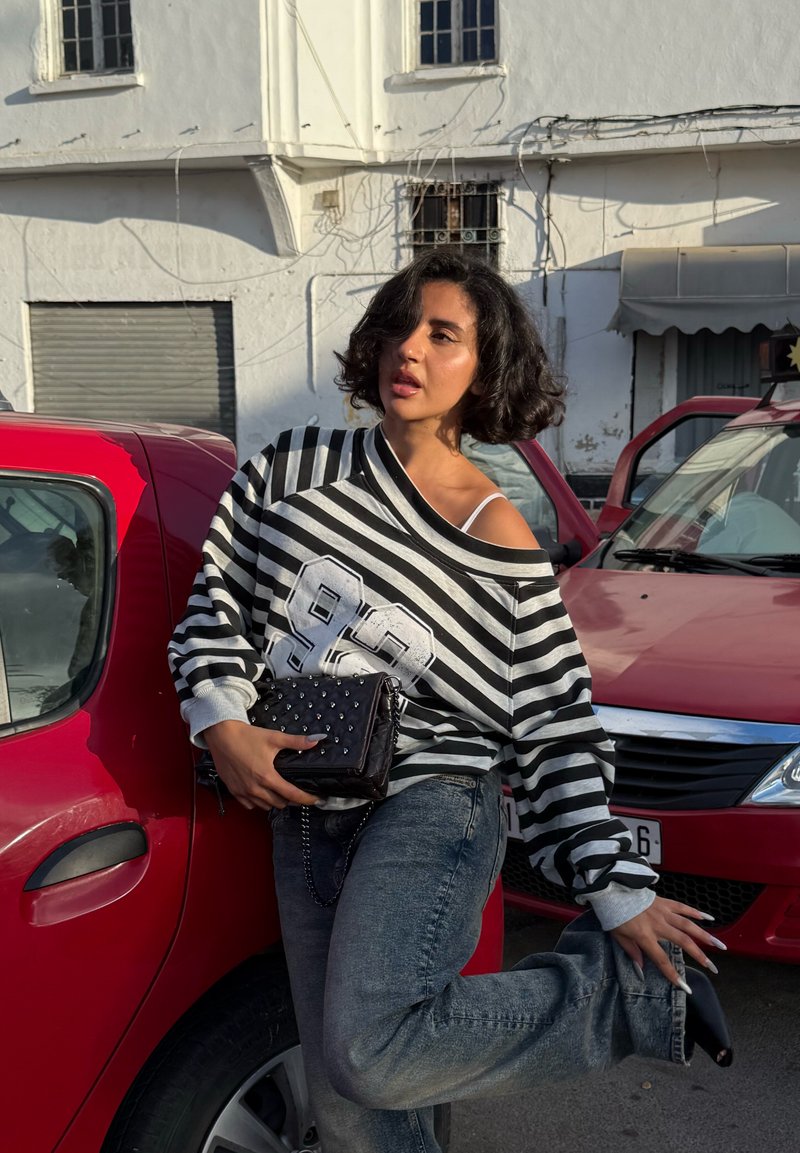 Schwarz-weiß gestreifter, übergroßer Pullover mit Zahlendetail. Dunkle Jeans und eine schwarze Tasche mit Nieten. Rotes Auto im Hintergrund.