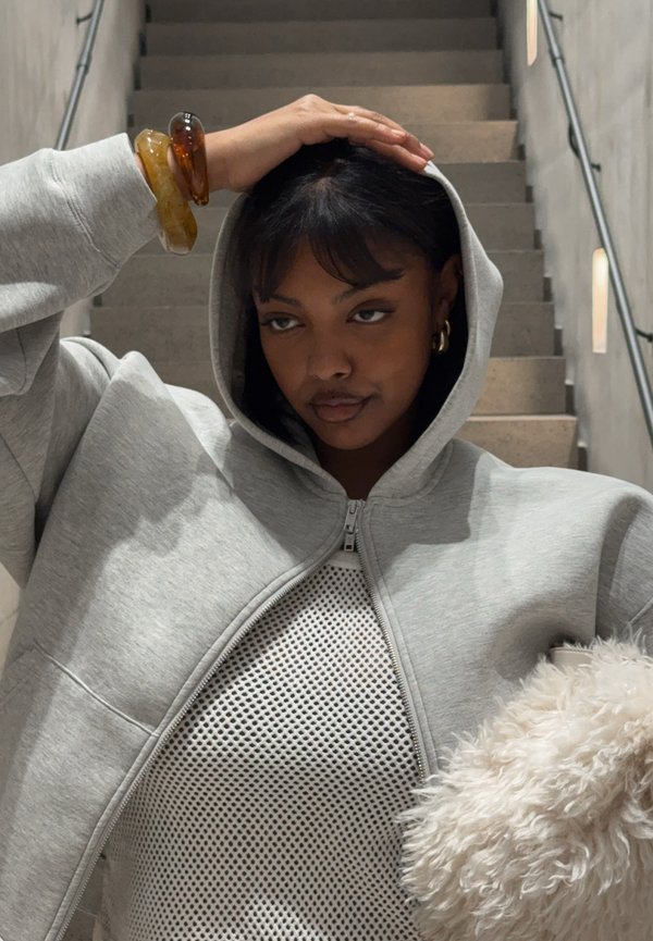 Sudadera con capucha gris con cremallera, capucha grande, material suave de algodón, bolsillos delanteros, lleva encima una camiseta de malla, complementada con un accesorio blanco y esponjoso.