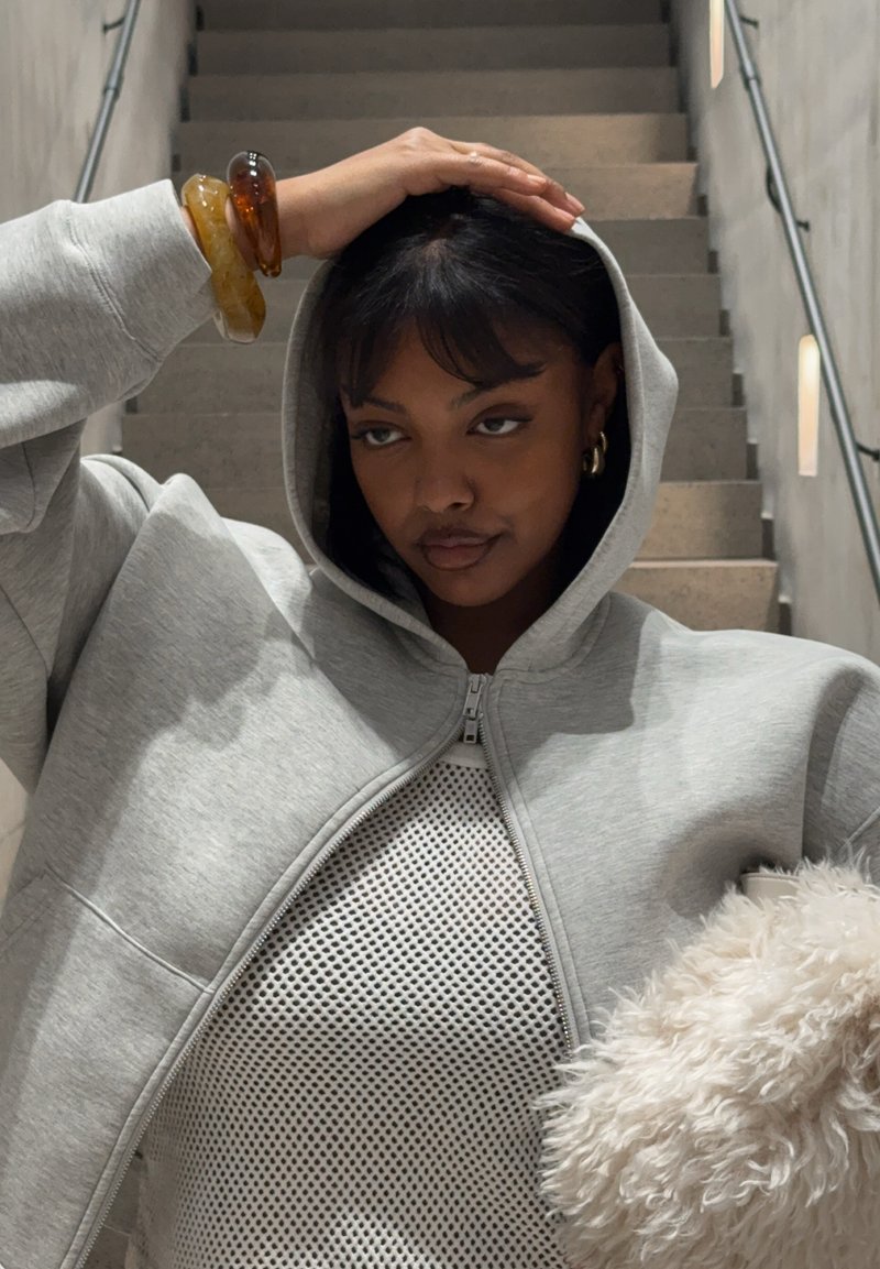 Grå hættetrøje med lynlås og stor hætte, blødt bomuldsstof, frontlommer, båret over en nettop, suppleret med en fluffet hvid accessory.