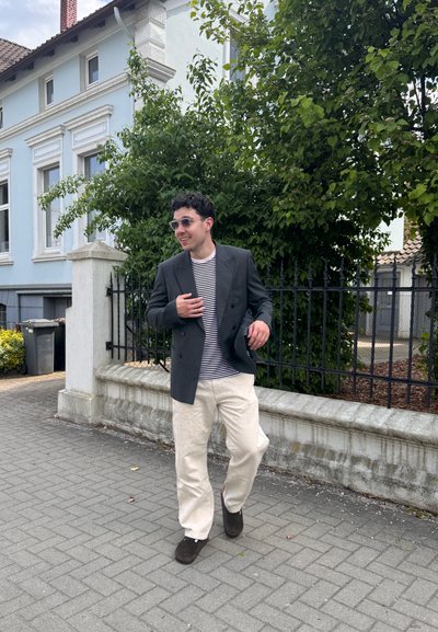 Hombre con gafas de sol, blazer oscuro, camisa a rayas y pantalones claros caminando por una acera junto a una pared blanca y árboles verdes.