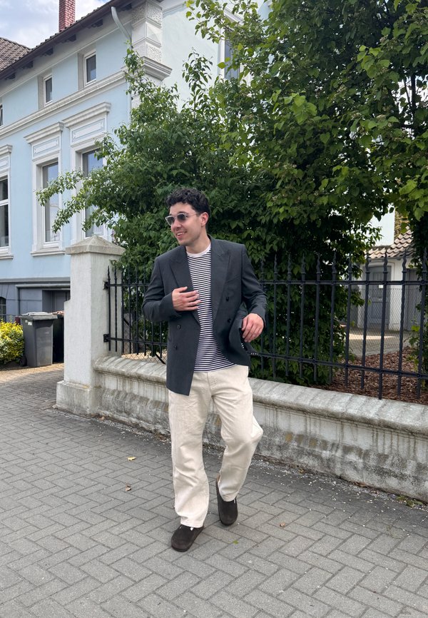 Homme portant des lunettes de soleil, un blazer foncé, une chemise rayée et un pantalon clair marchant sur un trottoir à côté d'un mur blanc et d'arbres verts.