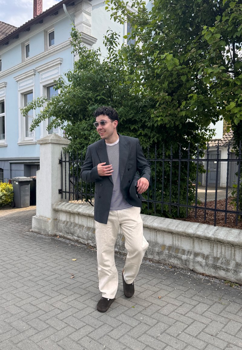 Homme portant des lunettes de soleil, un blazer sombre, une chemise rayée et un pantalon clair marchant sur un trottoir à côté d'un mur blanc et d’arbres verts.