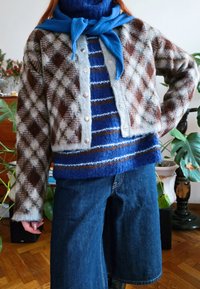 Άτομο που φοράει μπλε ριγέ πουλόβερ κάτω από καφέ καρό ζακέτα με κουμπιά, φαρδιά σκούρα μπλε τζιν παντελόνια και μπλε μαντήλι δεμένο γύρω από το λαιμό.