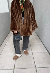 Καφέ ψεύτικη γούνα oversized παλτό, πράσινο κα patterned κασκόλ, γκρι παντελόνι σε φαρδιά γραμμή και καφέ παντόφλες με λευκή γούνα. Μαύρη τσάντα χειρός με αλυσίδα.