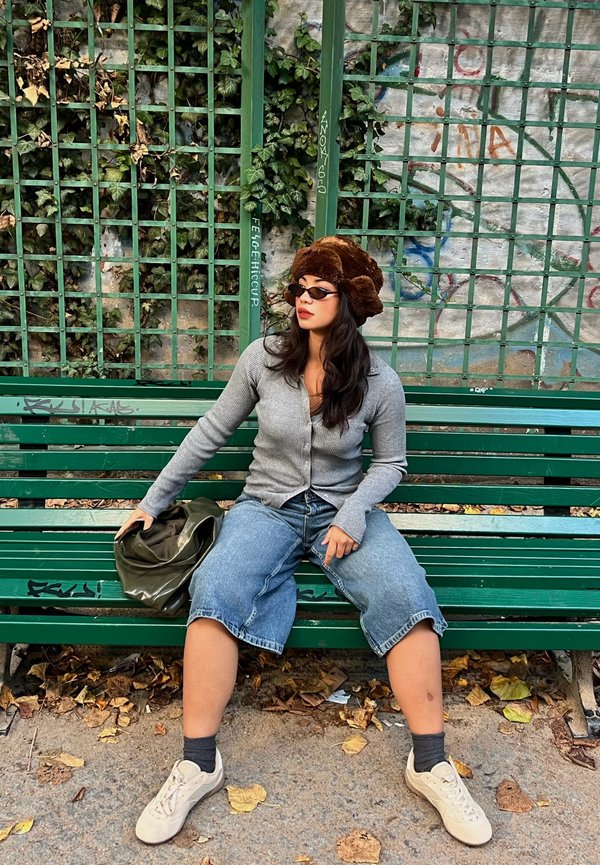 Grijze button-up cardigan, bruine nepbont hoed, denim culottes en witte sneakers. Zittend op een groene bank omringd door gevallen bladeren.