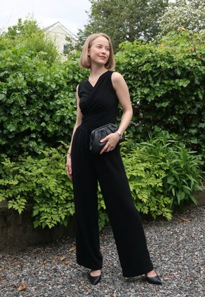 Mujer con mono negro sin mangas sostiene un bolso negro, de pie sobre grava con arbustos verdes y árboles al fondo.