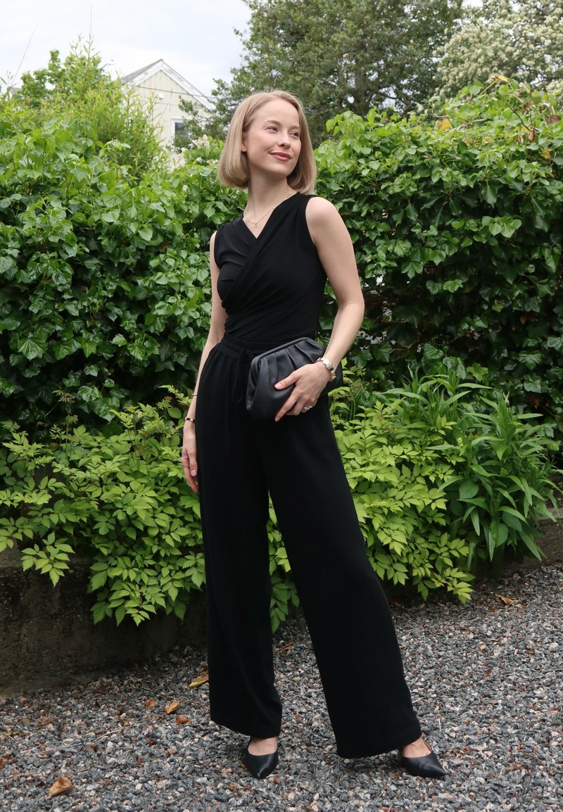 Femme en combinaison noire sans manches tenant une pochette noire, debout sur du gravier avec des buissons verts et des arbres en arrière-plan.