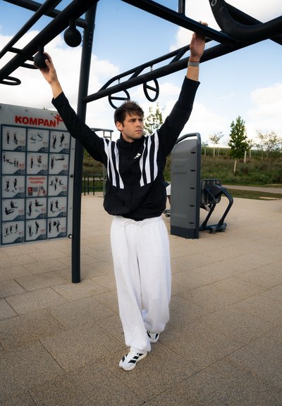 Una persona realiza un ejercicio colgado en una estructura de fitness exterior negra, vistiendo una chaqueta negra con rayas blancas y pantalones de chándal blancos.