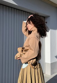 Veste beige surdimensionnée à col montant, associée à une chemise bleu clair et une longue jupe plissée kaki, tenant des chaussures et un sac beiges.
