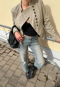 Persona con chaqueta beige con botones, top negro, vaqueros azul claro, botas negras y sosteniendo un bolso negro, de pie junto a una barandilla de metal al aire libre.