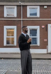 Černý pletený kardigan přes světle modrou košili, šedé široké kalhoty, stojící na dlažebních kostkách; v pozadí několik oken cihlové budovy.