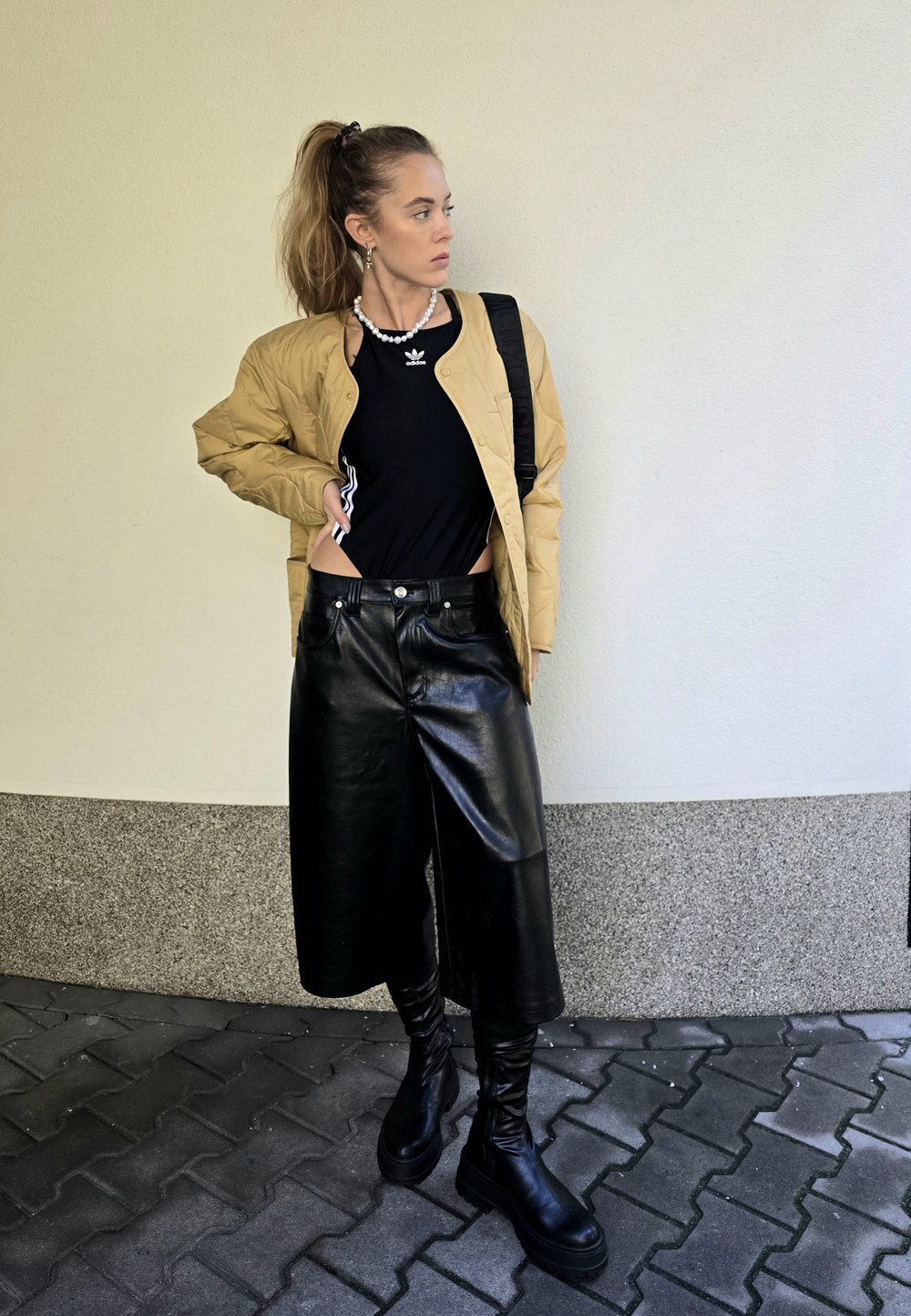 Ταν πλισέ μπομπερ τζάκετ πάνω από μαύρη μπλούζα, σε συνδυασμό με φαρδιές μαύρες δερμάτινες κολάν και μπότες μέχρι το γόνατο σε μαύρο χρώμα. Λευκό φόντο τοίχου.