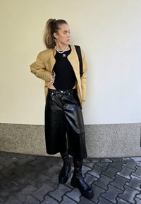 Ταν πλισέ μπομπερ τζάκετ πάνω από μαύρη μπλούζα, σε συνδυασμό με φαρδιές μαύρες δερμάτινες κολάν και μπότες μέχρι το γόνατο σε μαύρο χρώμα. Λευκό φόντο τοίχου.