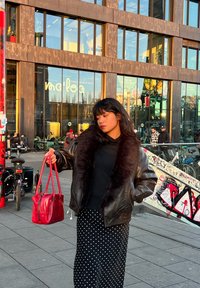 Chaqueta de cuero negra con cuello de piel, suéter negro, falda de lunares y bolso rojo. Entorno urbano con edificios de cristal al fondo.