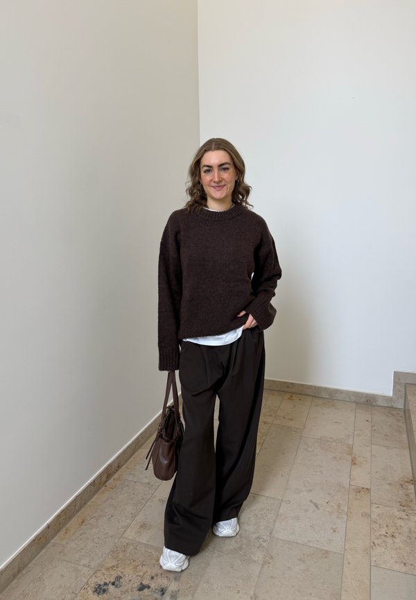 Femme aux cheveux bouclés portant un pull marron foncé, un pantalon noir ample, des baskets blanches, portant un sac marron, debout à l'intérieur contre un mur uni.