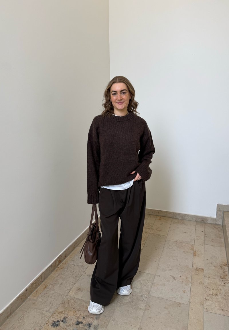 Frau mit lockigem Haar, die einen dunkelbraunen Pullover, lockere schwarze Hose, weiße Turnschuhe trägt, eine braune Tasche bei sich hat und drinnen vor einer schlichten Wand steht.