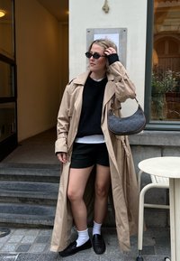 Trench-coat beige, pull noir, short noir, chaussettes blanches et mocassins foncés. Porte un sac à main noir à motifs. Debout sur des marches en pierre.