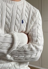 Personne portant un pull en maille torsadée blanc avec un logo de joueur de polo brodé en bleu, bras croisés devant un fond neutre.