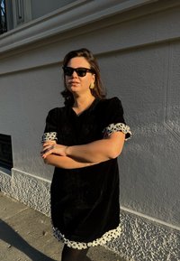 Zwarte fluwelen jurk met pofmouwen en een witte stippenrand bij de manchetten en zoom. Gecombineerd met een zwarte zonnebril en gouden oorbellen.