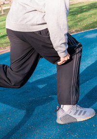 Joggers en polaire noire avec trois bandes latérales, associés à des chaussures de sport grises avec une semelle texturée et un design à lacets, sur un sol bleu.