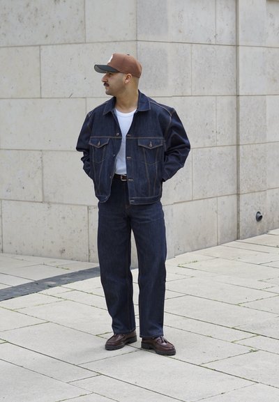 Chaqueta de mezclilla oscura y pantalones con una camiseta blanca, gorra marrón y zapatos marrones oscuros, de pie sobre una superficie pavimentada contra una pared de piedra.