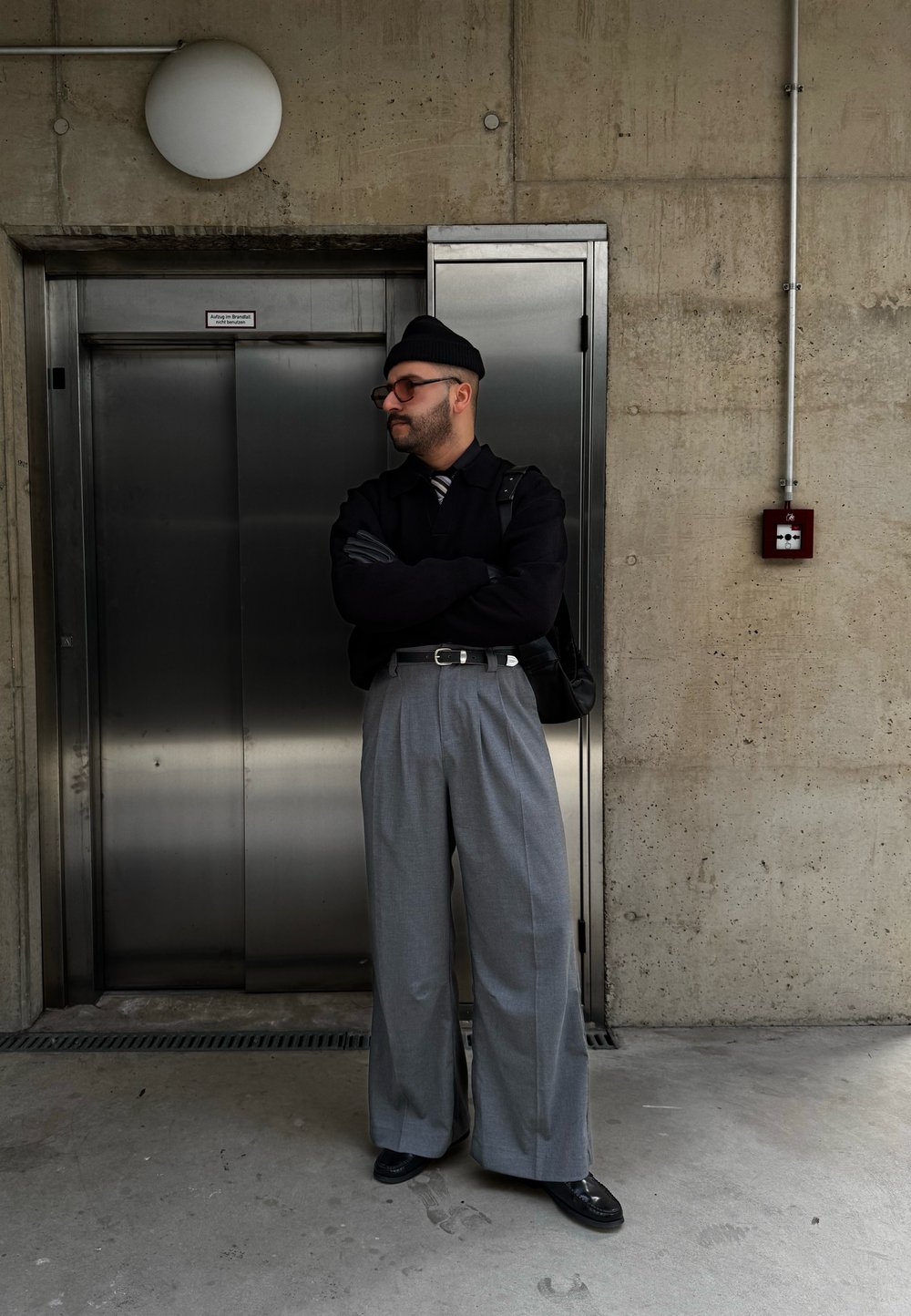 Γκρι παντελόνι φαρδιάς γραμμής με ψηλή μέση, συνδυασμένο με μαύρο πουλόβερ και γραβάτα. Αξεσουάρ περιλαμβάνουν γυαλιά και μαύρ ο σκουφί. Ανελκυστήρας στο φόντο.