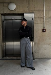 Γκρι παντελόνι φαρδιάς γραμμής με ψηλή μέση, συνδυασμένο με μαύρο πουλόβερ και γραβάτα. Αξεσουάρ περιλαμβάνουν γυαλιά και μαύρο σκουφί. Ανελκυστήρας στο φόντο.