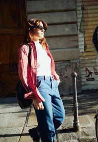 Donna con occhiali da sole, camicia a righe rosse, top bianco e jeans blu, che si trova su una strada soleggiata vicino a un muro di pietra e a un palo di metallo.