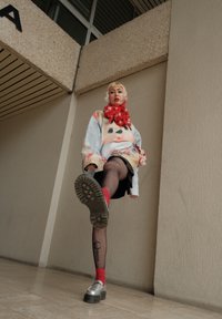 Maglione lavorato a maglia con un design di faccia di bambola, gonna nera, calze fino al ginocchio rosse e scarpe con plateaux argentate. Lo sfondo presenta pareti in cemento testurizzato.