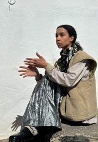 Brun kordflorväst över en rutmönstrad skjorta, kombinerad med en glänsande silverkjol. Svarta skor med dekorativa nitar. Blommig scarf runt halsen.