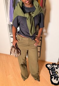 Personne portant un pantalon vert olive et un t-shirt noir avec un pull vert noué sur les épaules, tenant un sac à main marron à motifs à l'intérieur.