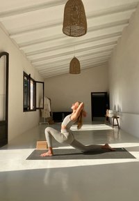 Osoba w jasnoszarym stroju sportowym wykonuje pozę jogi na ciemnej macie w minimalistycznym pokoju z białym sufitem i plecionymi lampami.