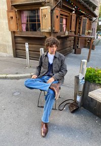 Καθημερινό outfit που περιλαμβάνει ένα γκρι μπουφάν, μπλε πλεκτό γιλέκο, ανοιχτό μπλε τζιν και καφέ δερμάτινα μποτάκια. Σκηνικό έξω από μια ρουστίκ ξύλινη κατασκευή.