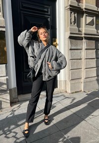 Gestepptes graues Bomberjacke, schwarze Hose und besetzte schwarze Ballerinas. Steht gegen eine Tür mit goldenem Beschlag, Sonnenlicht wirft Schatten.