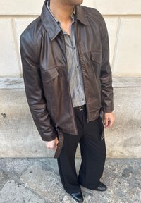 Mężczyzna w ciemnobrązowej skórzanej kurtce, pasiastym koszulowym zapiętym na guziki, czarnych spodniach i czarnych butach stoi na kamiennej nawierzchni przy beżowej ścianie.