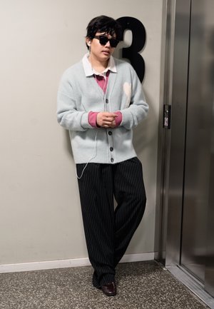 Persona con gafas de sol, cárdigan gris, pantalones a rayas, escuchando auriculares, apoyada en una pared numerada con el número 3 cerca de las puertas del ascensor.