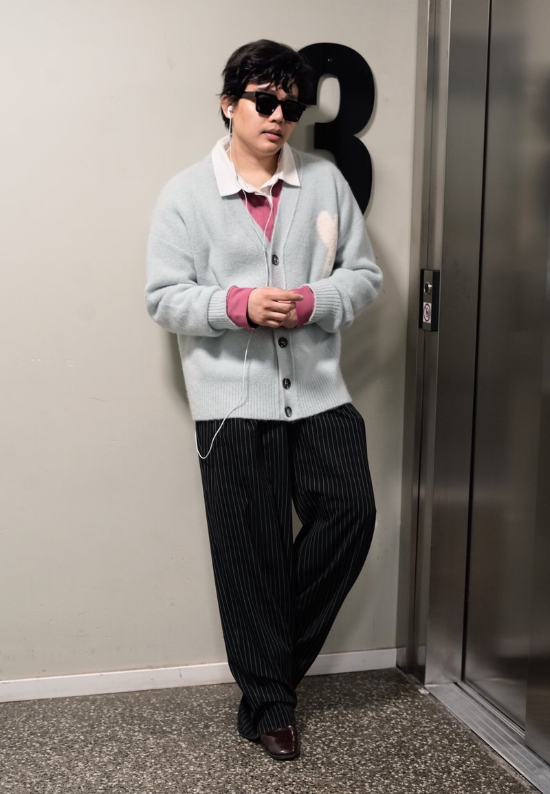 Personne portant des lunettes de soleil, un cardigan gris, un pantalon rayé, écoutant des écouteurs, appuyée contre un mur numéroté 3 près des portes de l'ascenseur.