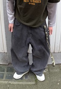 Baggy sorte jeans med en tekstureret finish og sidelomme, sammen med en mørkegrøn t-shirt med orange tekst, båret over en hvid langærmet skjorte.