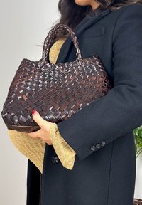 Femme en manteau noir tenant un grand sac à main en cuir tressé brun foncé avec des poignées tressées, par-dessus un pull beige.