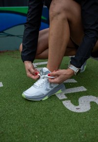 Zapatilla deportiva blanca con acentos grises y suela de un verde brillante. Persona atándose los cordones, arrodillada sobre un césped verde con marcas blancas.