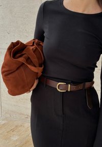 Brūna suede zīmuļa soma ar melnu pieguļošu augšdaļu un brūnu ādas jostu ar zelta sprādzi, fona tekstūrā.