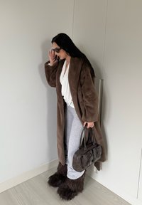 Manteau en fausse fourrure marron porté sur un haut blanc, un pantalon de survêtement gris et des bottes marron en fourrure. Les accessoires comprennent des lunettes de soleil foncées et un sac à main noir.